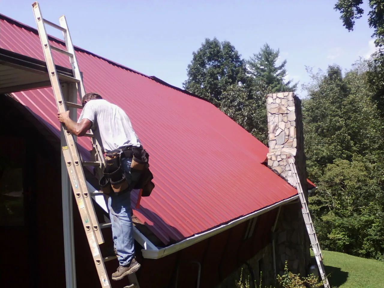 Roofer on a ladder beside a red metal roof and stone chimney — Soffit & Fascia Repair by Roofing Brothers in Quinton